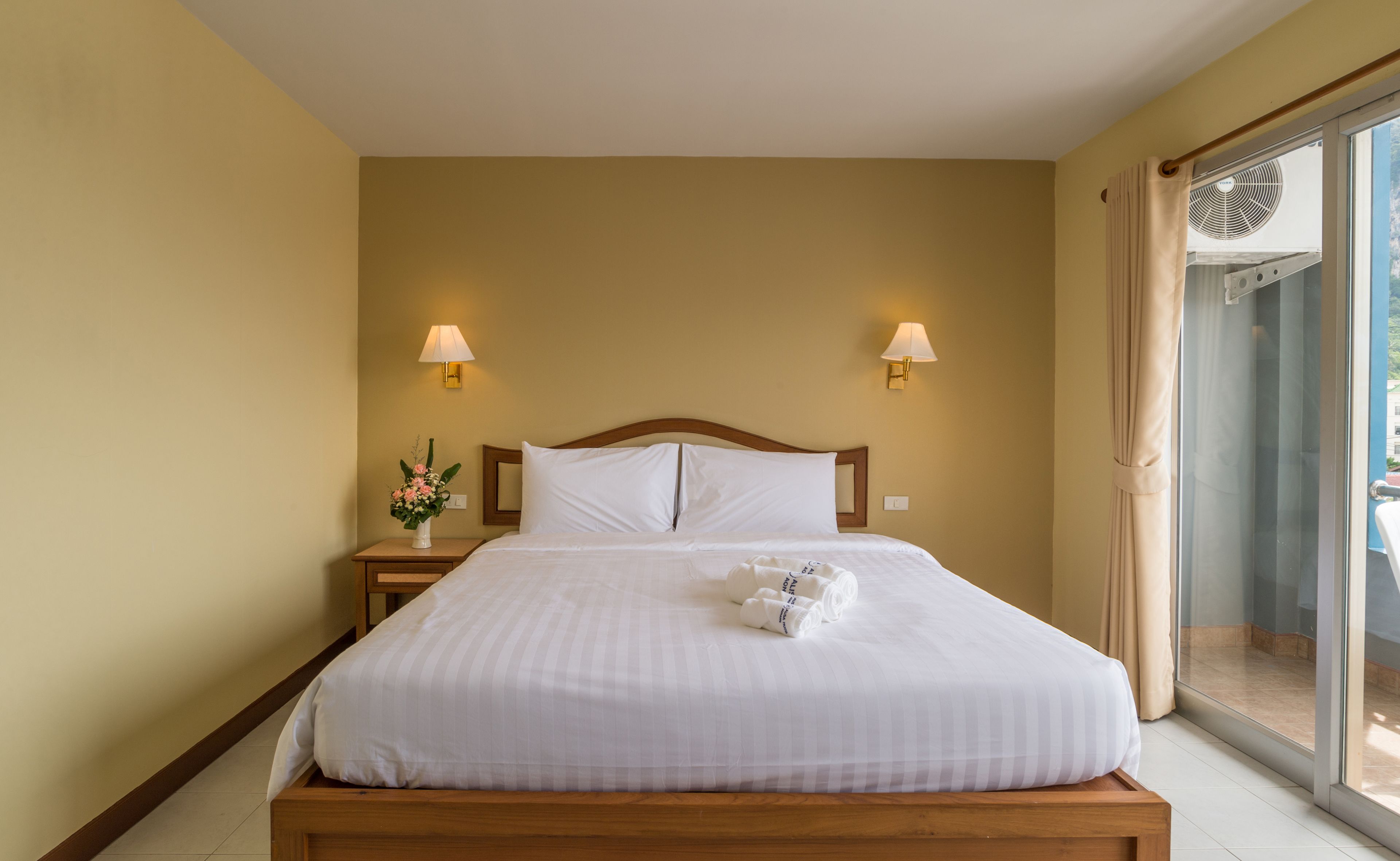 A hotel room with a bed, two bedside lamps, a nightstand with a vase of flowers, and a view of the outside through a glass door.