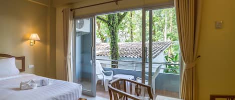 A hotel room with a bed, a table with a book, a chair, and a balcony with a view of greenery and a tiled roof.