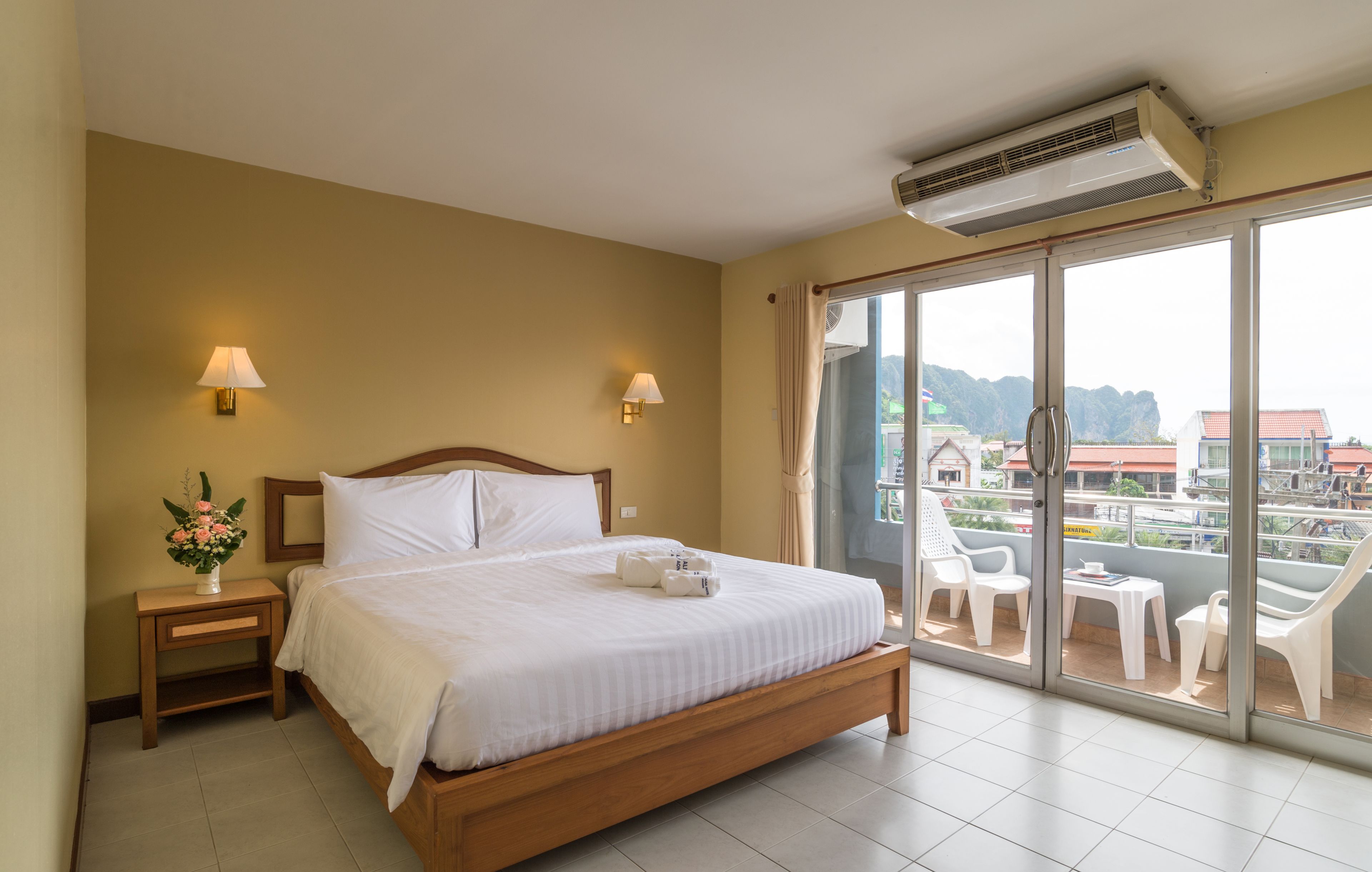 Une chambre d’hôtel avec un lit, des lampes de chevet, une petite table ornée d’un vase de fleurs et un balcon avec des chaises et une table.