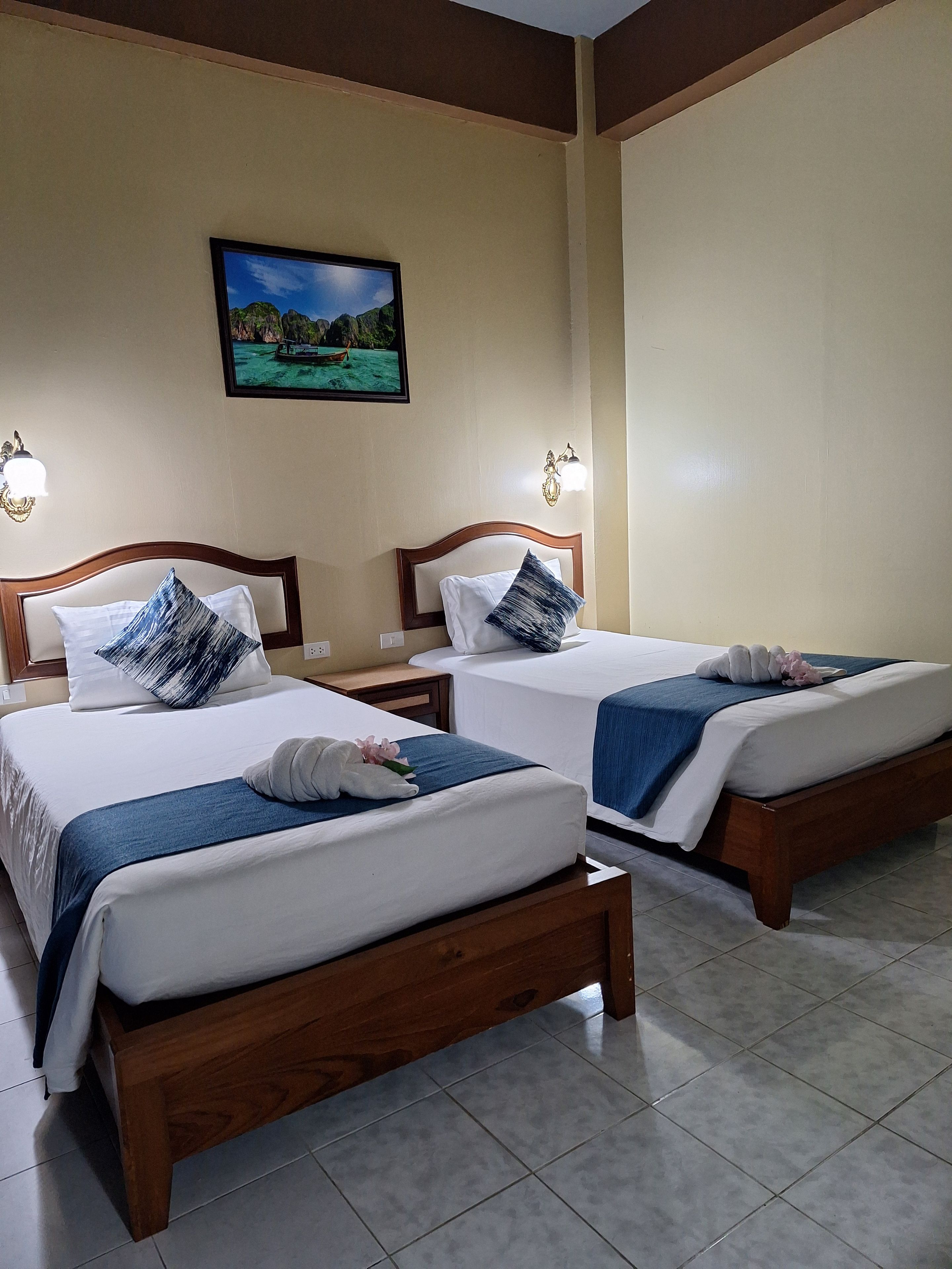 Une chambre d’hôtel avec deux lits en bois, chacun doté d’un matelas blanc, d’une couverture bleue et de coussins décoratifs. Des appliques murales et un tableau encadré ornent le mur au-dessus des lits.