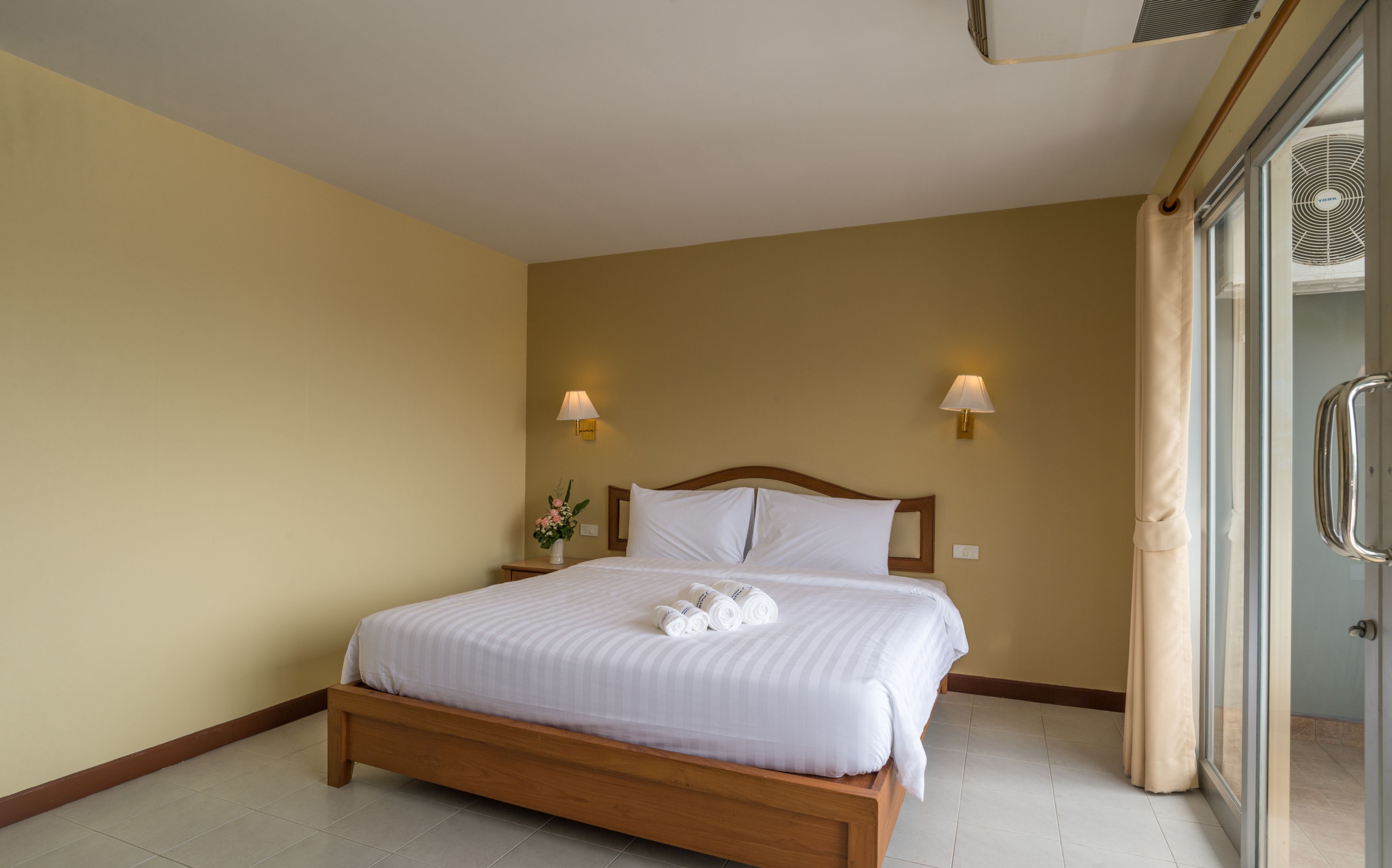 Ein Hotelzimmer mit einem hölzernen Bett, zwei Wandlampen, einer Vase mit Blumen und einer Glastür zum Badezimmer.
