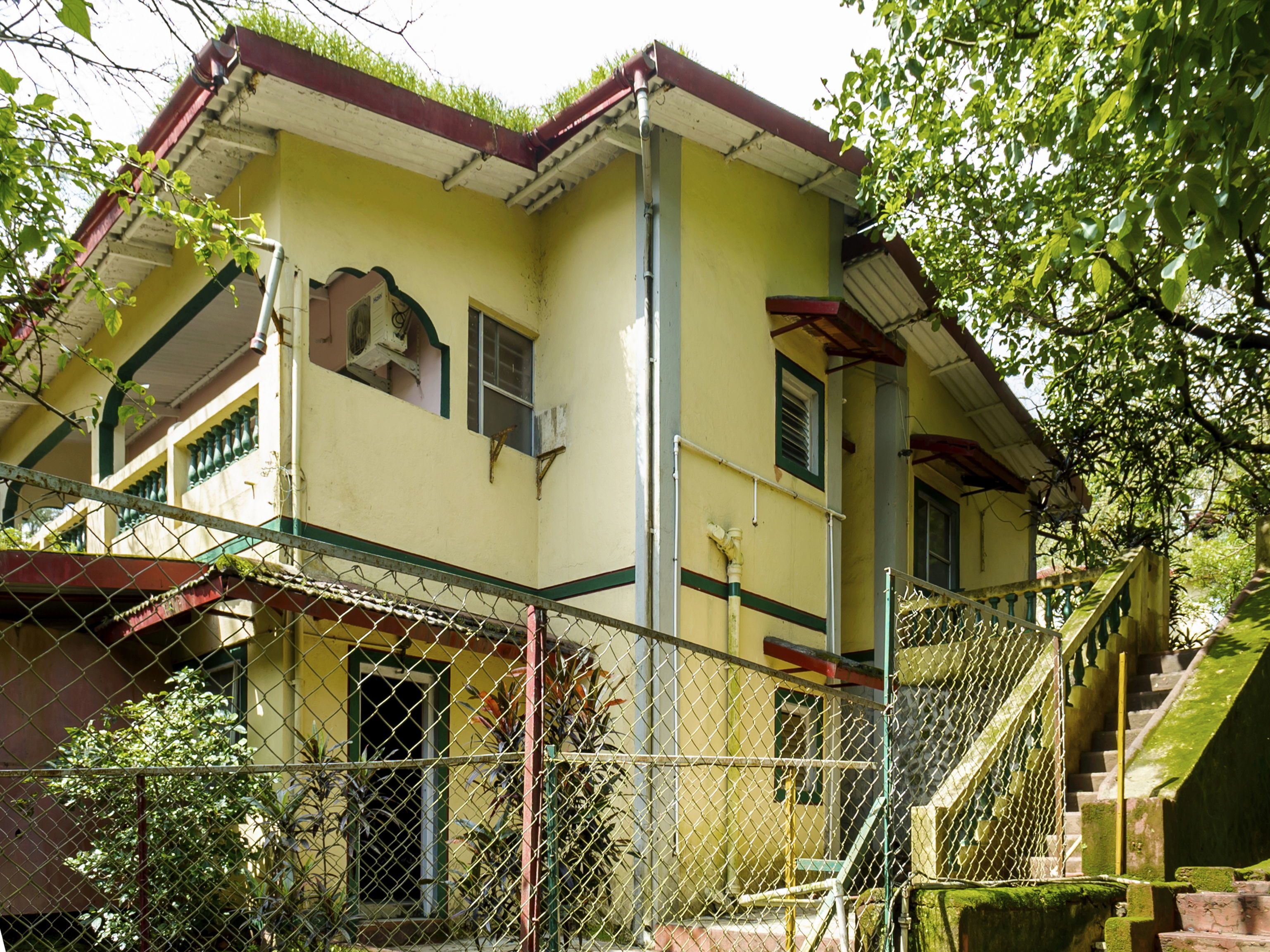 A two-story building with a yellow facade, green window frames, and a red roof. There is a staircase leading to the upper floor, and a chain-link fence surrounds the property.