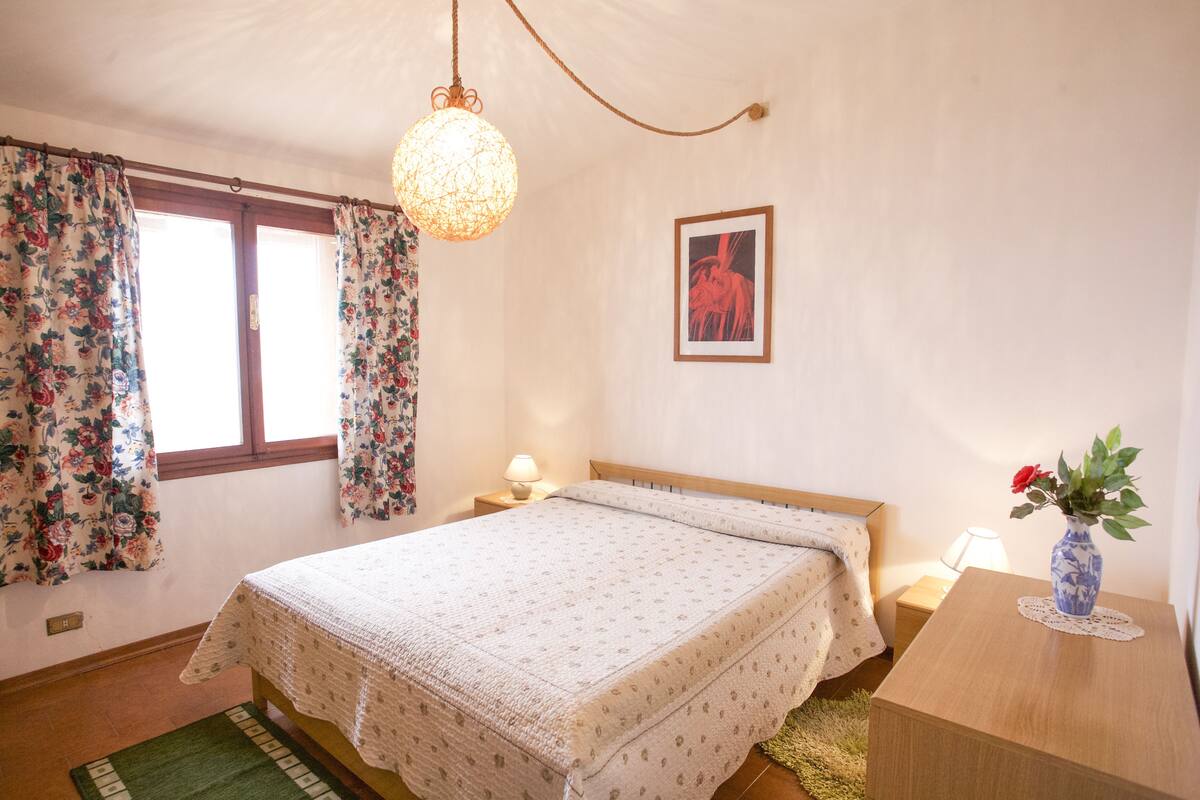 Une chambre à coucher avec un lit, des rideaux, un luminaire suspendu, une table de chevet avec une lampe et un vase rempli de fleurs.