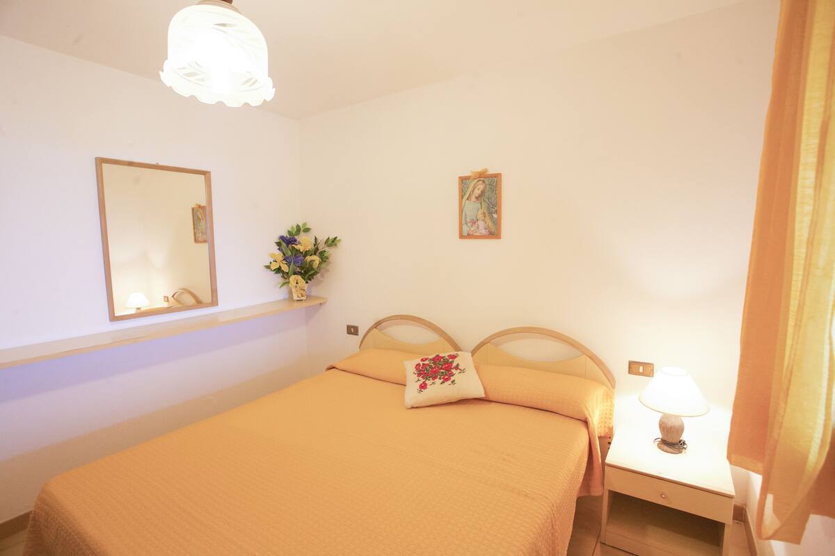 Une chambre à coucher avec un lit, une table de chevet avec une lampe, un miroir, un tableau au mur et un vase avec des fleurs.