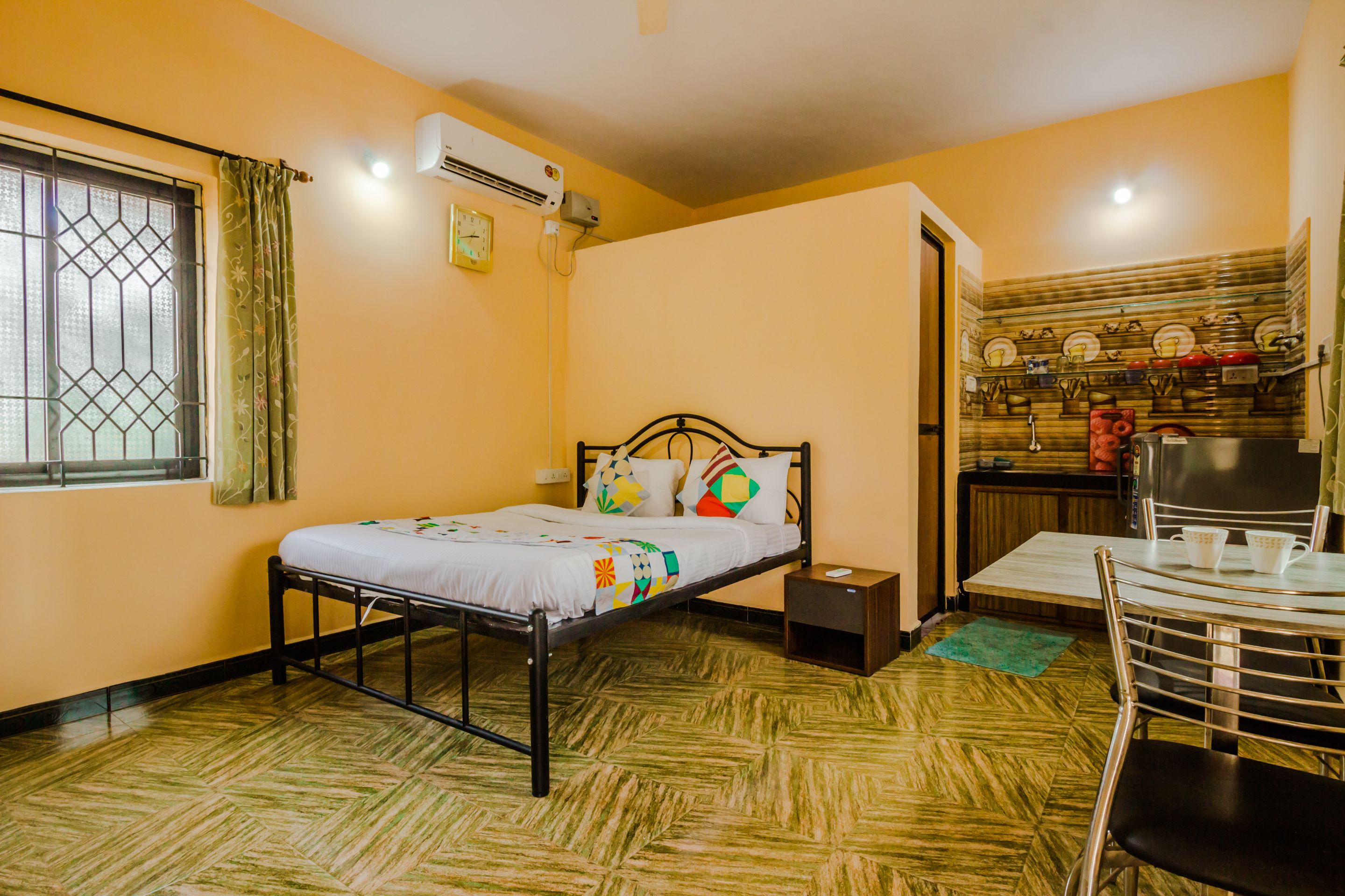A room with a bed, a small table, and a chair. There is a window with a curtain, an air conditioning unit, and a wall-mounted shelf with dishes.