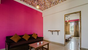 A room with a dark brown sofa against a bright pink wall, a wooden coffee table, and a mirror reflecting a bed with a window view. - OYO 22730 Home Pool View 1BHK Vagator Beach (Vagator)