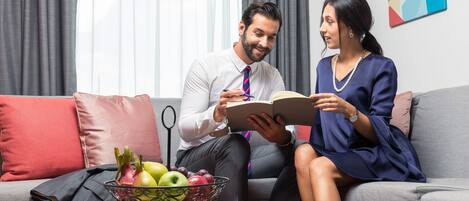 A man and a woman sitting on a grey sofa with a bowl of fruit on a stand in front of them.