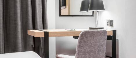 Una habitación de hotel moderna con un escritorio de madera, una silla tapizada en gris y una lámpara de pie negra.