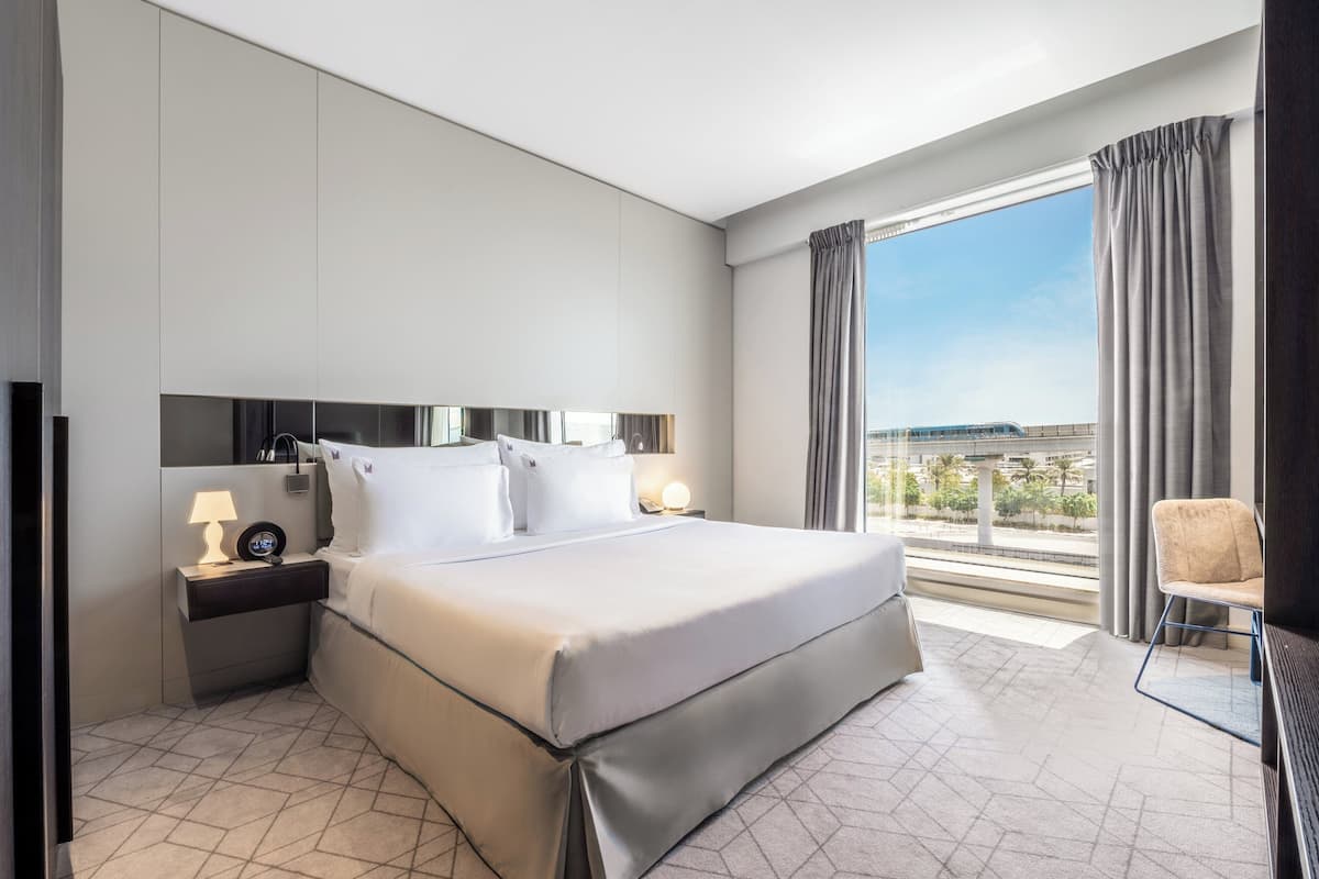 Ein modernes Hotelzimmer mit einem großen Bett, einem Nachttisch mit Lampe, einem Sessel und Blick auf die Stadt durch große Fenster.
