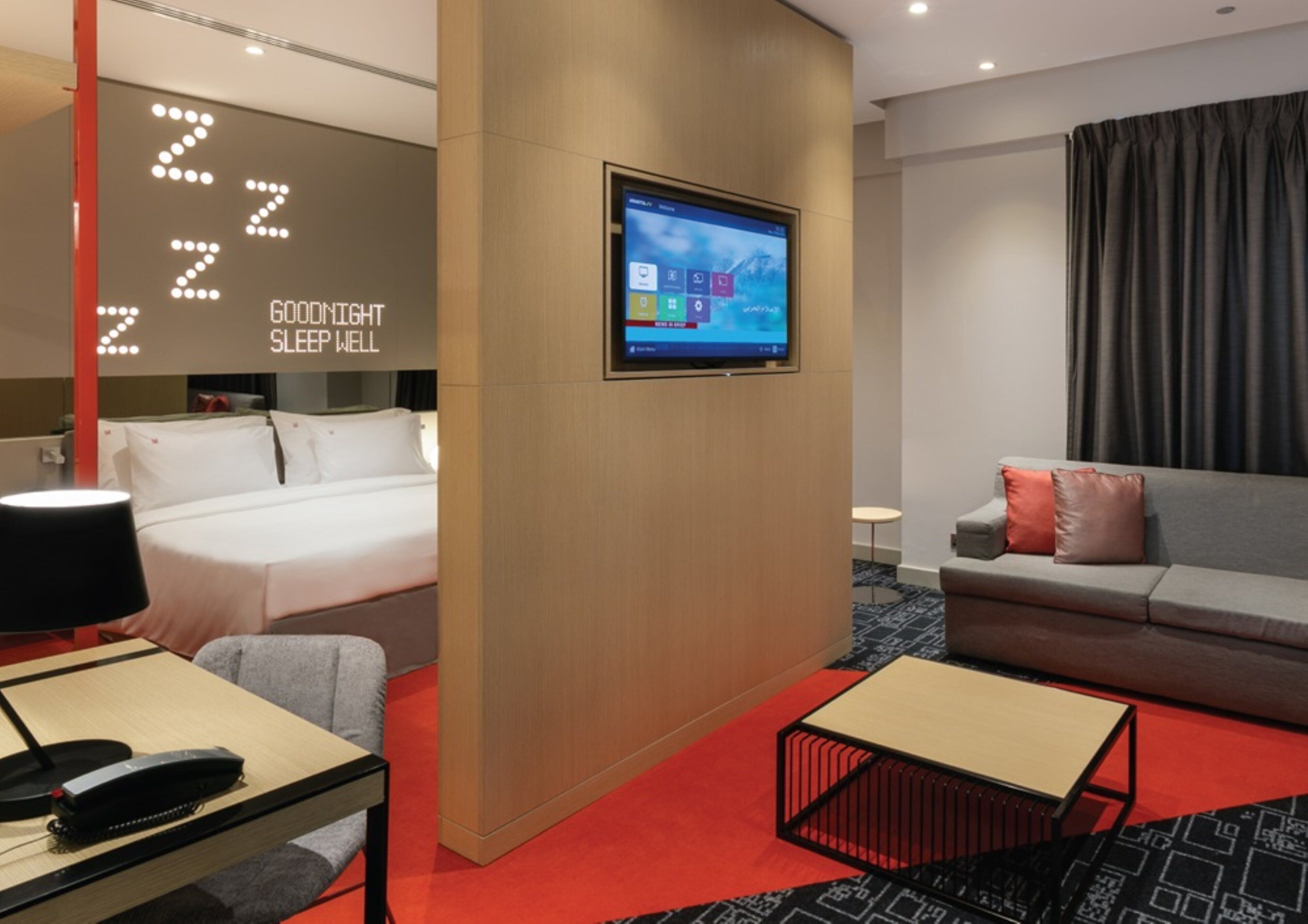 Ein Hotelzimmer mit einem Bett, einem an der Wand befestigten Fernseher, einer Couch und einem kleinen Tisch mit Lampe.