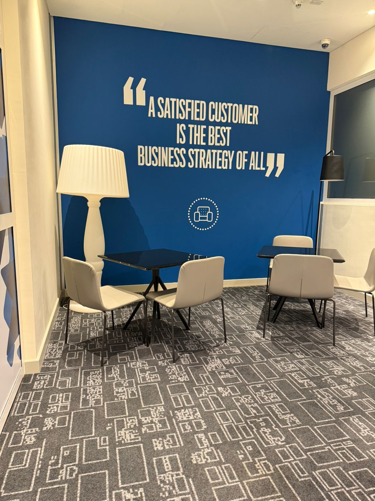 A room with a blue wall featuring a quote about customer satisfaction, a table with chairs, a lamp, and a patterned carpet.