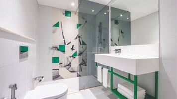 A modern bathroom with a white toilet, a glass-enclosed shower, and a white sink with a green shelf.