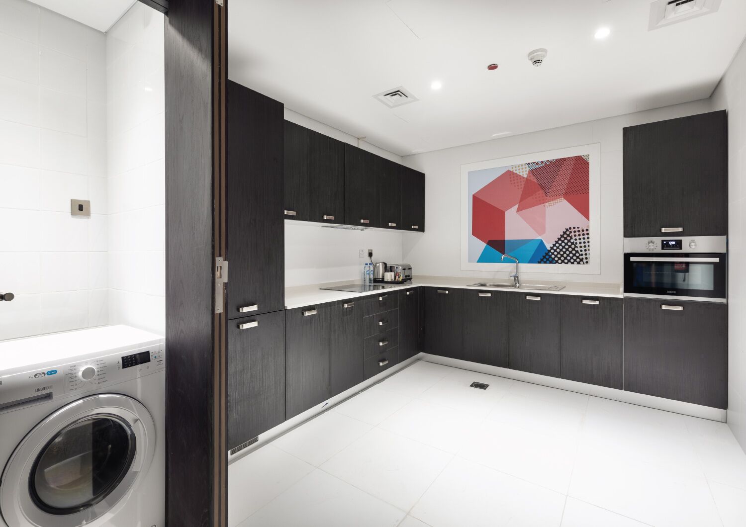 A modern kitchen with dark cabinetry, a built-in washing machine, and a large abstract painting on the wall.