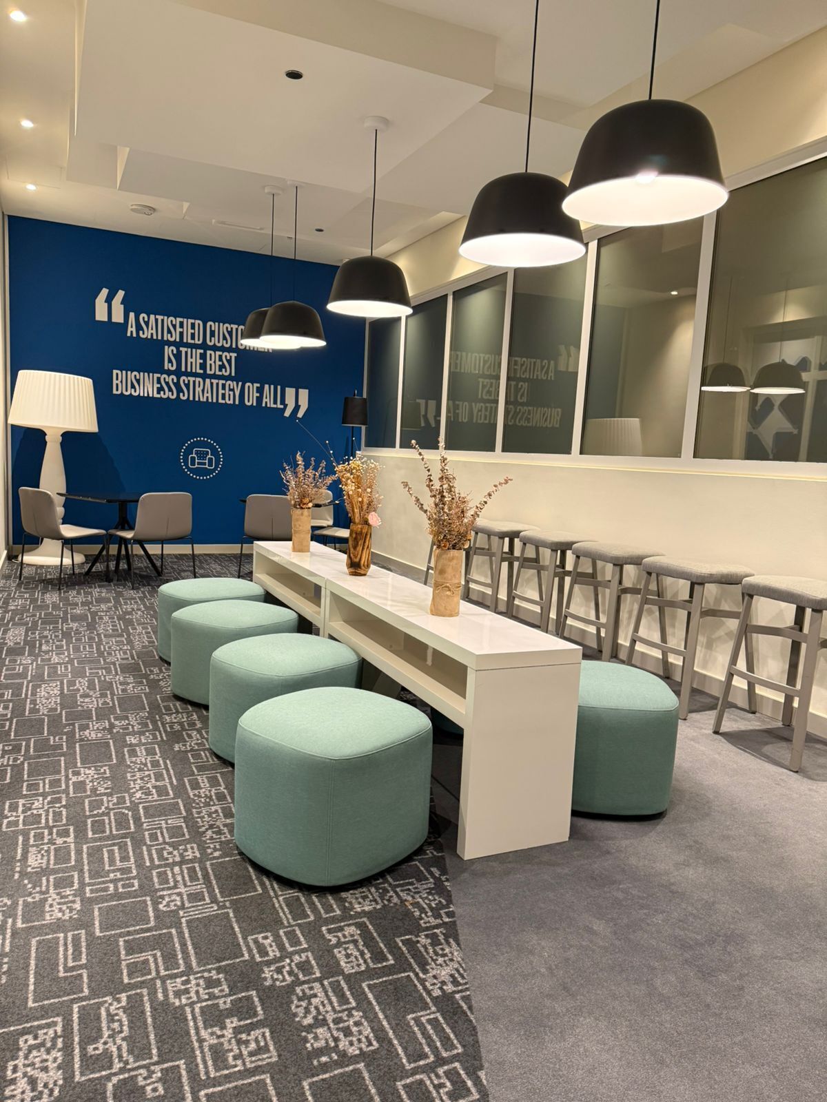 Un espacio de oficina moderno con una larga mesa blanca, poufs color turquesa y lámparas colgantes negras.