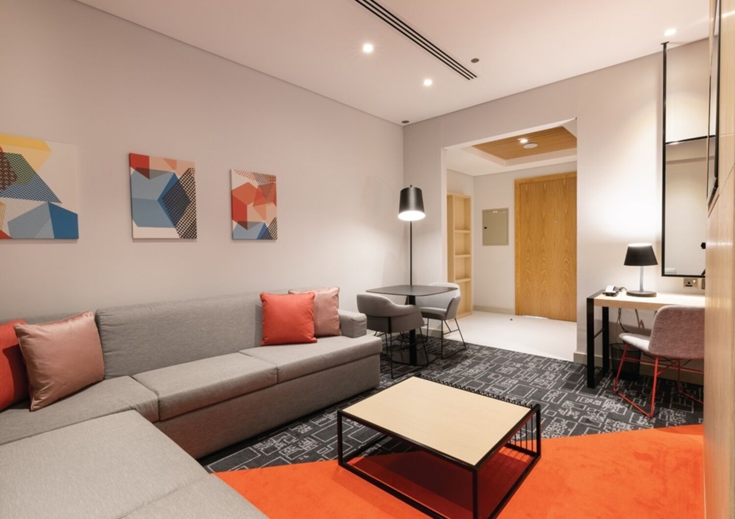 A modern hotel room with a grey sofa, a small round table, a desk with a chair, and abstract wall art.
