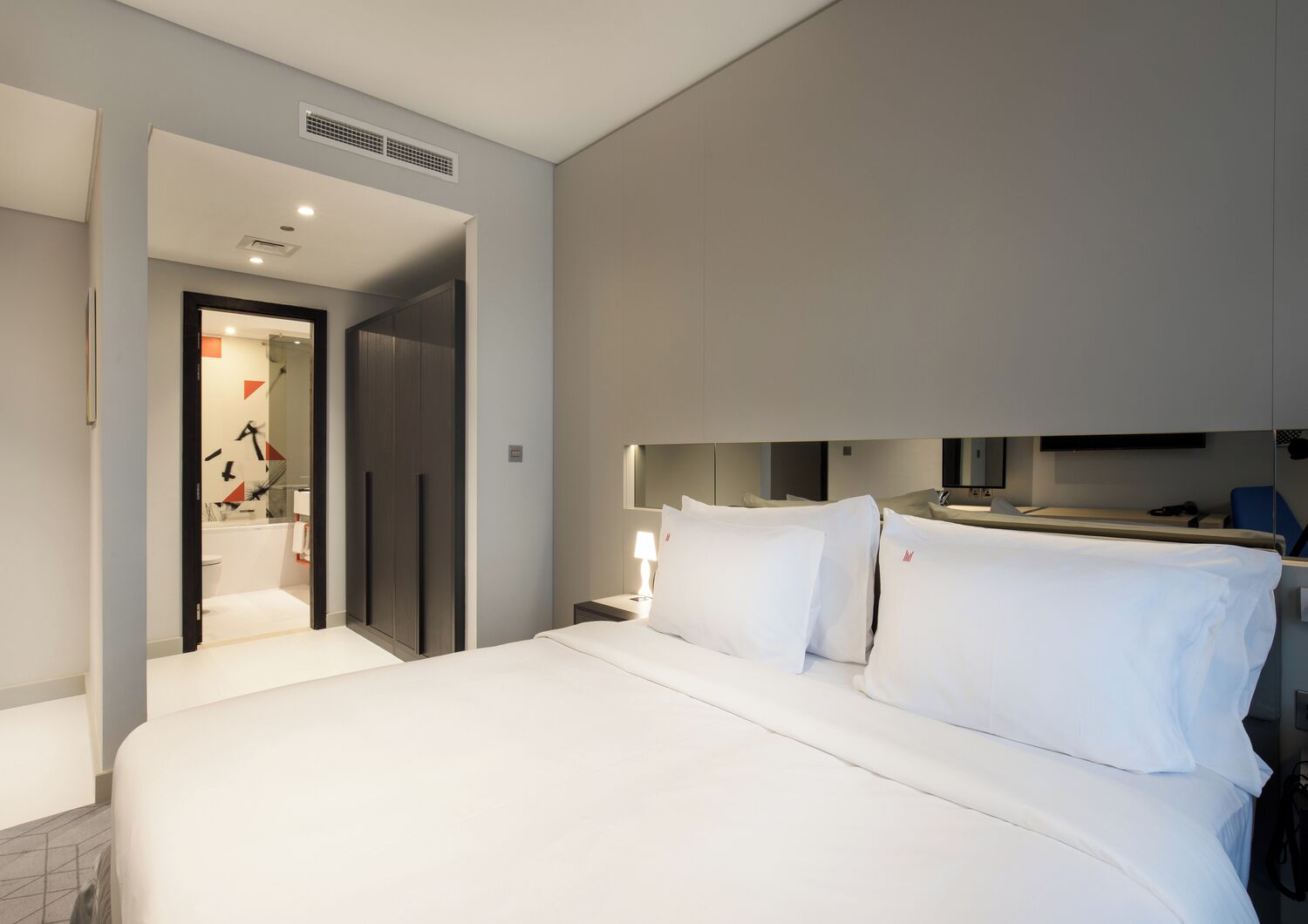 A hotel room with a large bed, a desk with a chair, a mirror, and a bathroom visible through an open door.