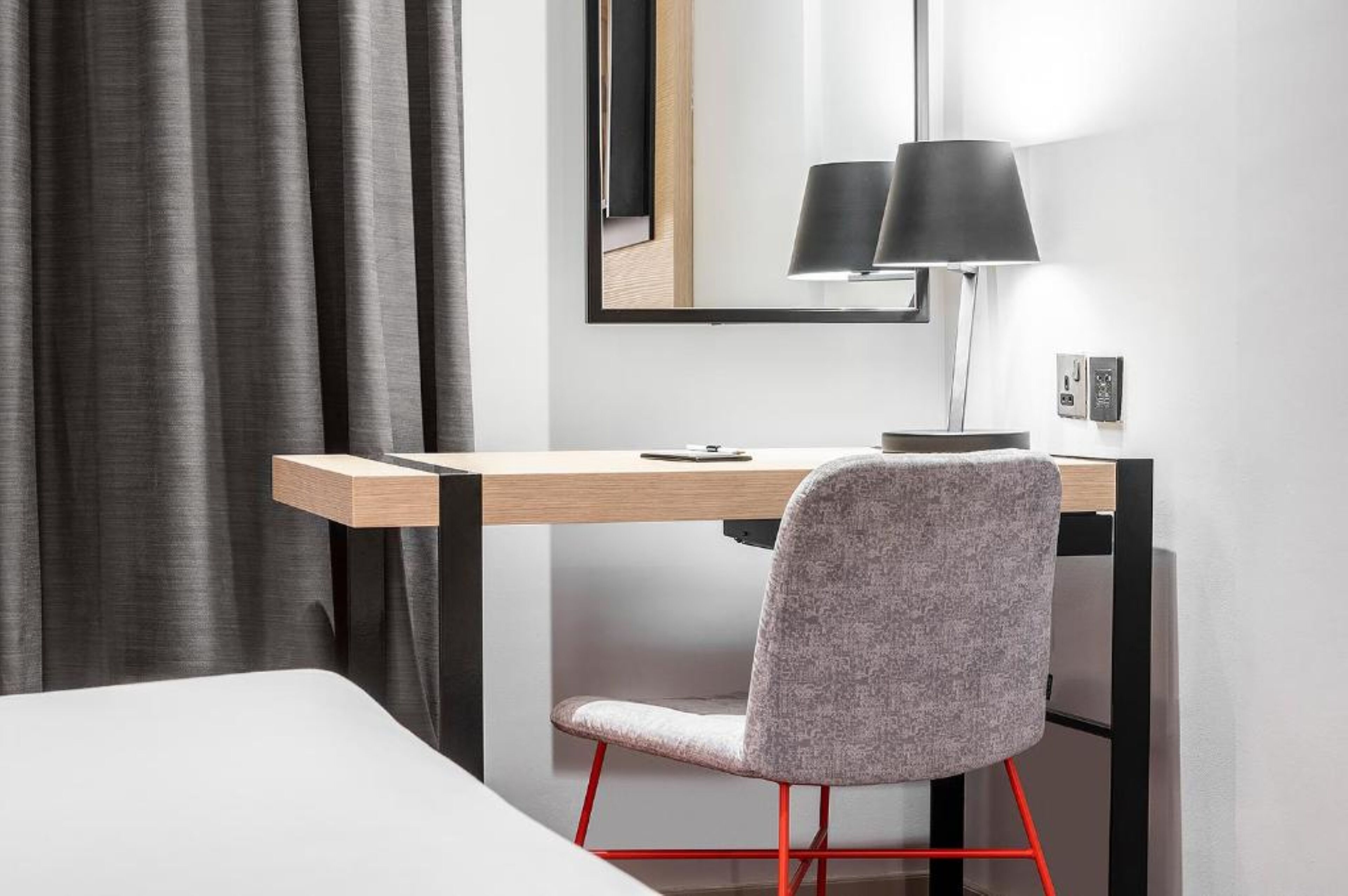 A modern hotel room with a wooden desk, a grey upholstered chair, and a black lamp.