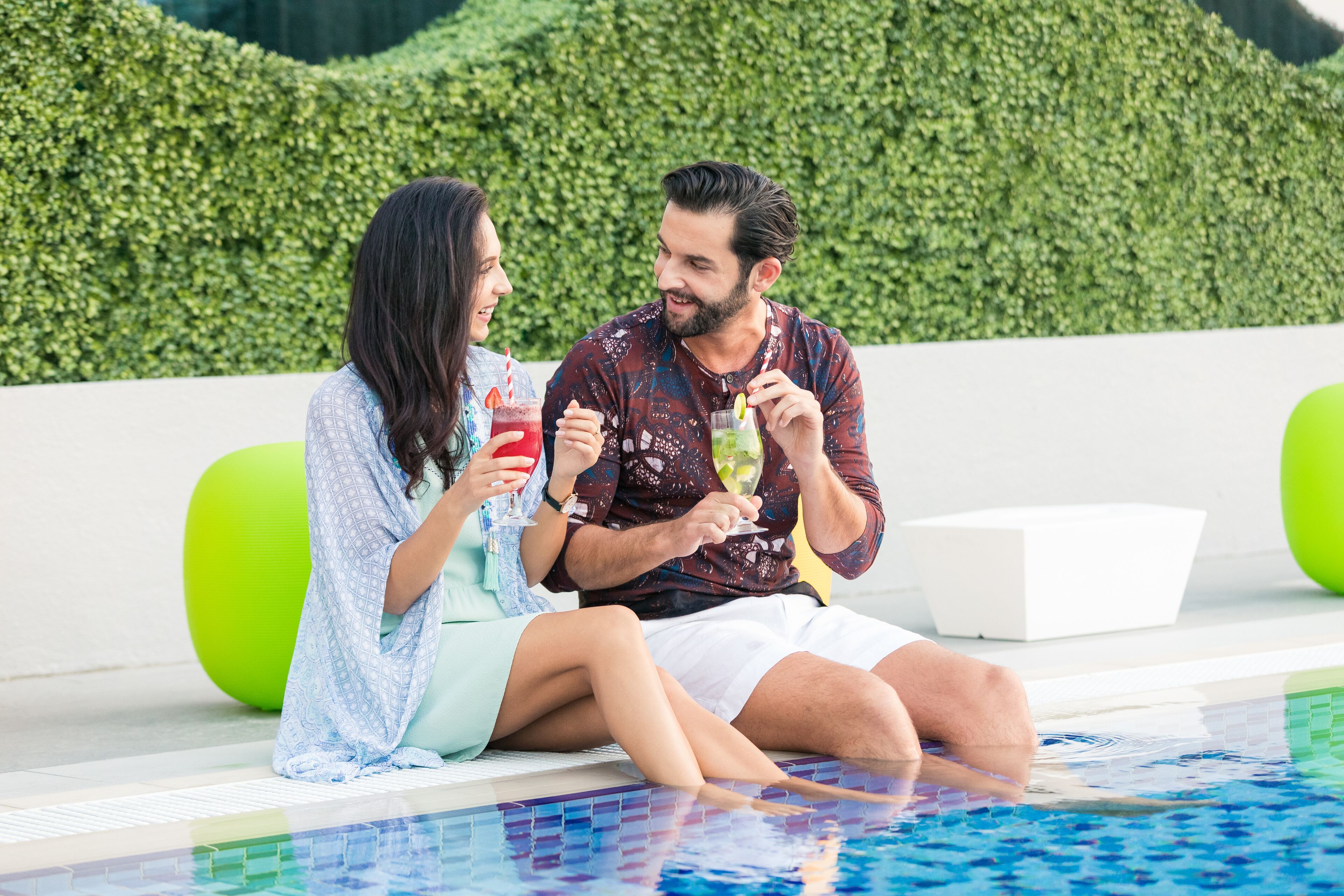 Un couple est assis au bord d’une piscine, des boissons à la main, entouré de verdure.