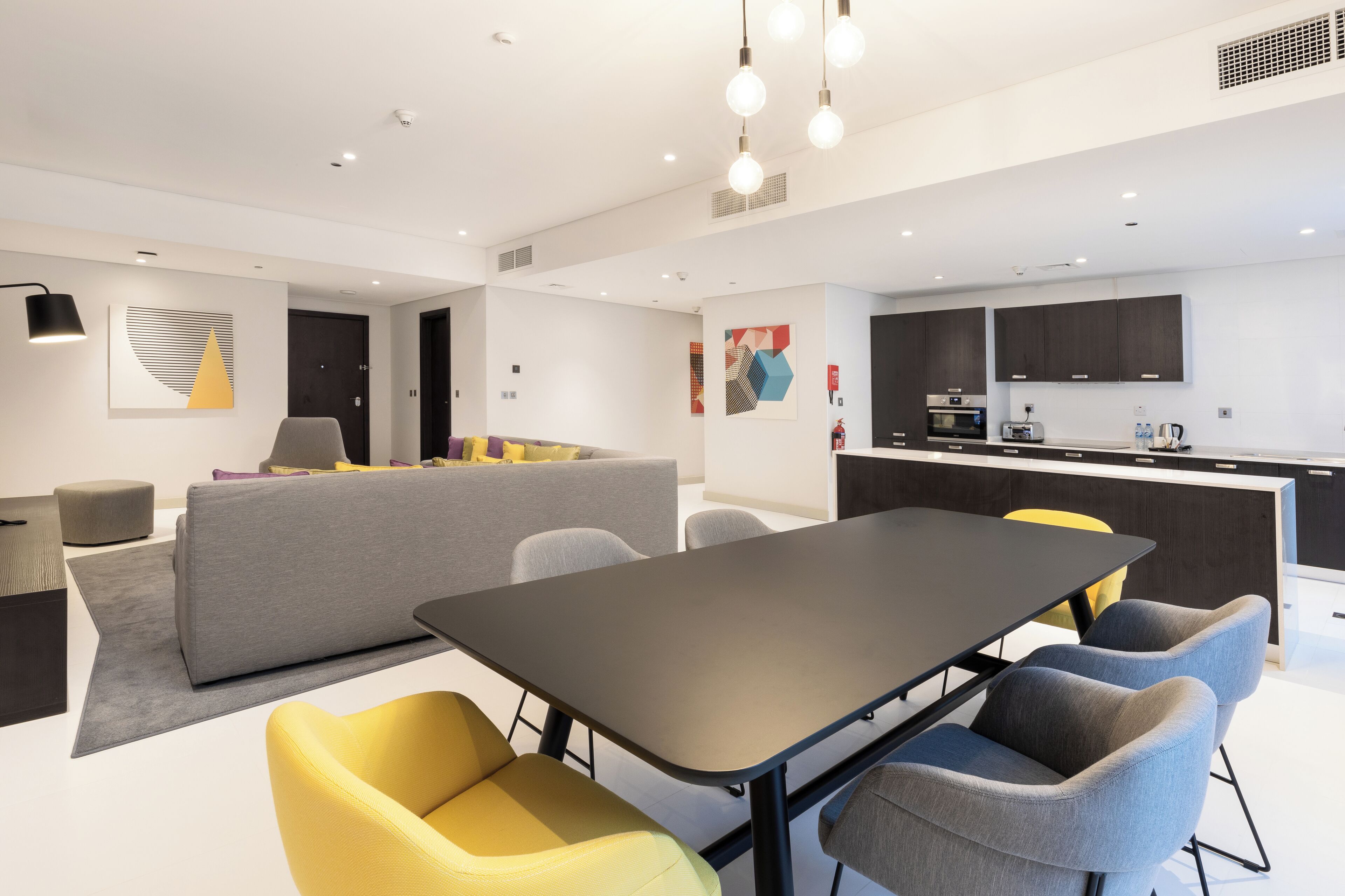 A modern living room with a sofa, a dining table, and chairs. There is a kitchen area with cabinets and appliances.