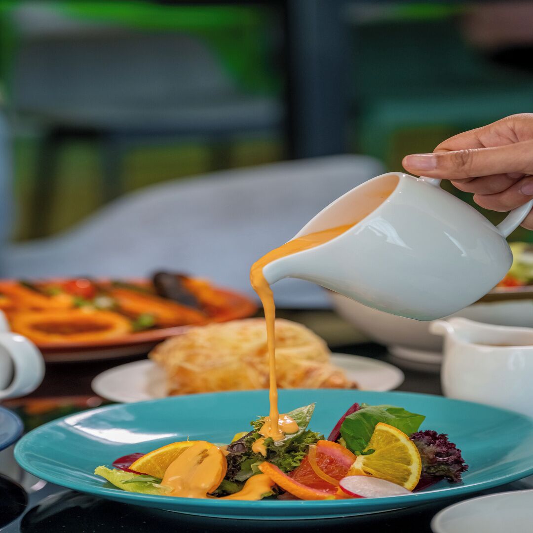 A person pouring a creamy sauce over a salad with various vegetables and fruits.