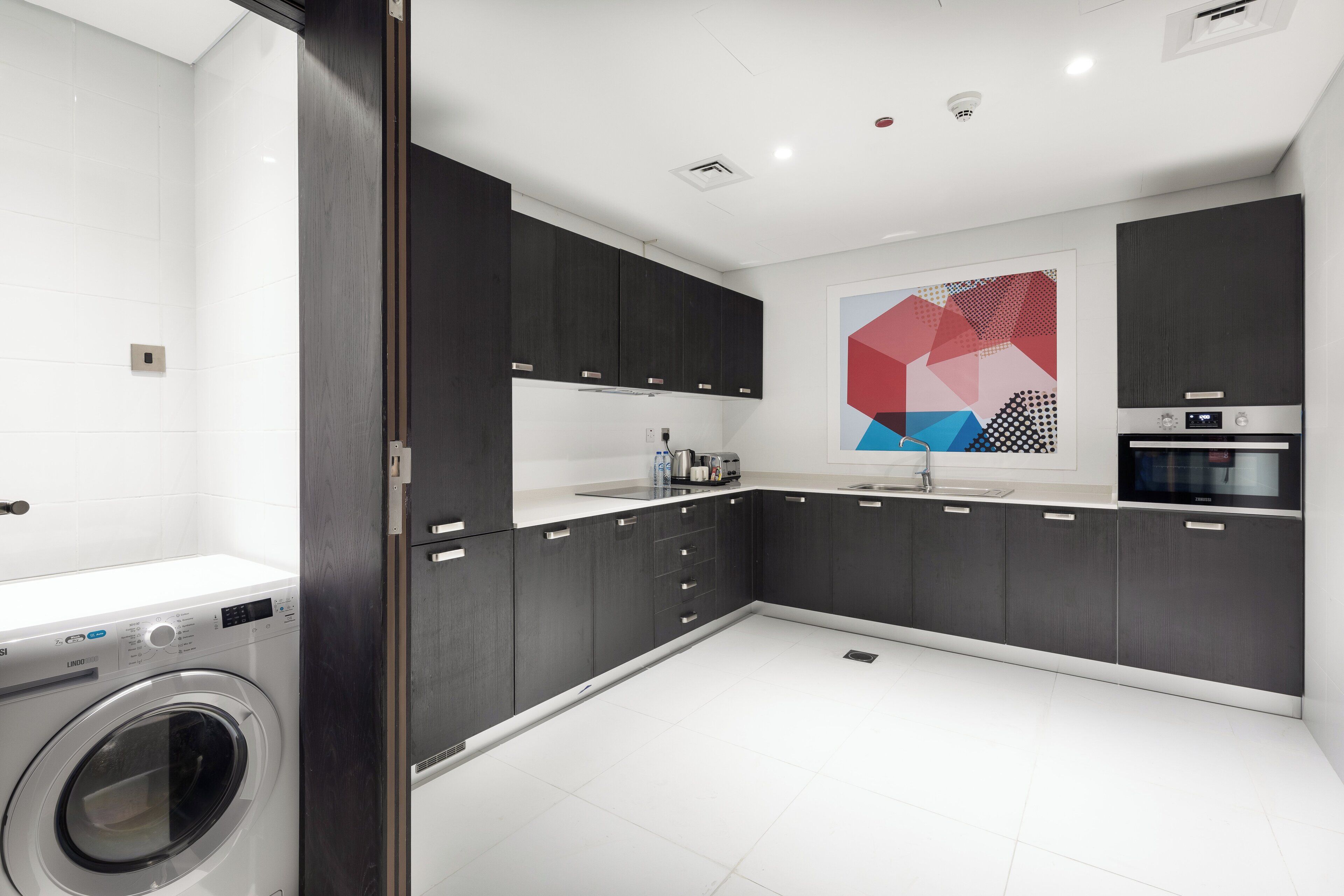 A modern kitchen with dark cabinetry, a built-in washing machine, and a geometric art piece on the wall.