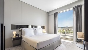 Quarto de hotel moderno com uma cama grande, uma mesa de cabeceira com abajur, vista da cidade através de grandes janelas e uma cadeira junto à janela.