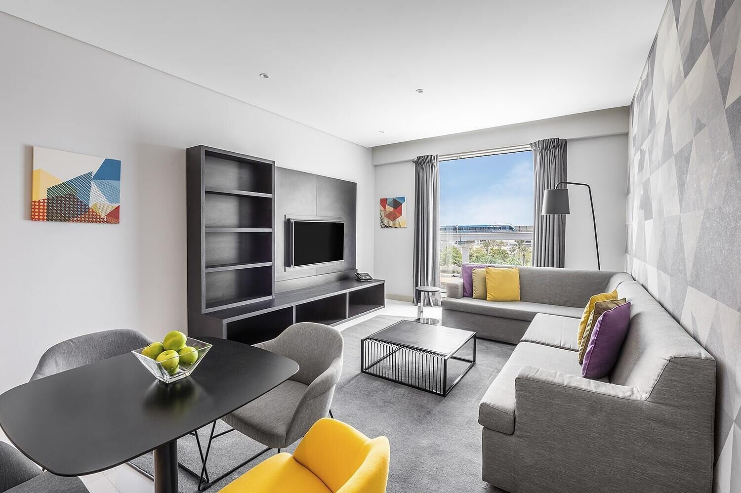 A modern living room with a grey sofa, a dining table with chairs, a TV mounted on a wall, and artwork on the walls.