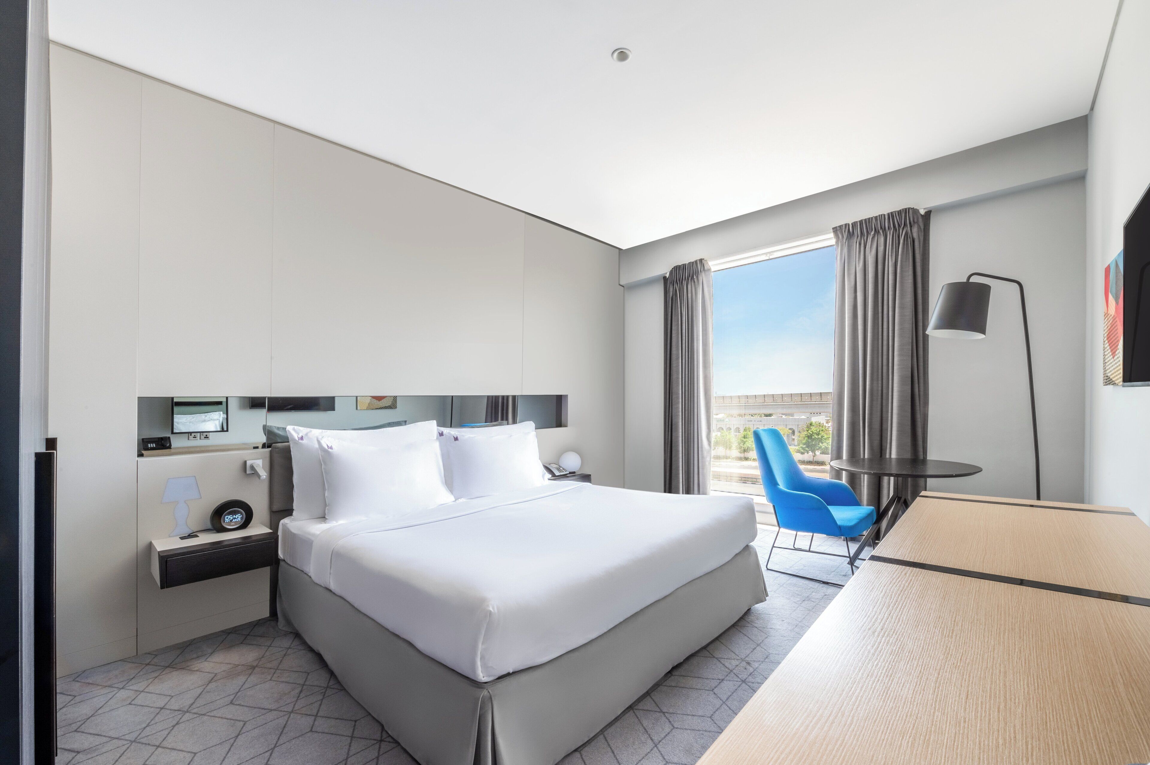 A modern hotel room with a large bed, a desk, a chair, a TV, and a window with curtains.