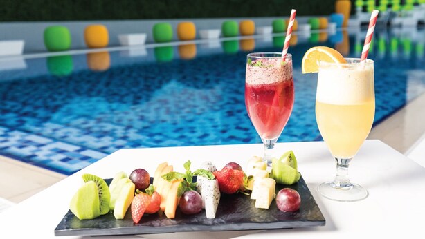Escena a la piscina amb un plat de fruita i dues begudes, una vermella i una groga, amb glaçons.