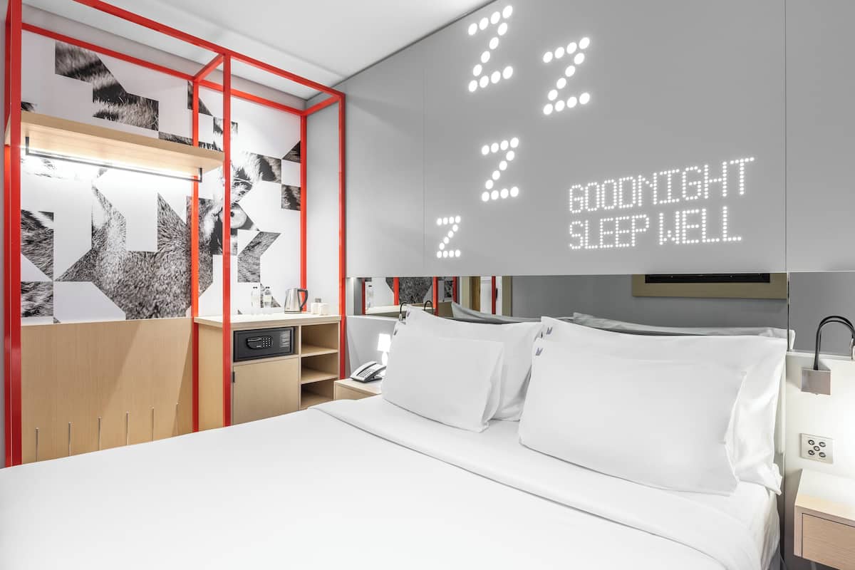 Ein modernes Hotelzimmer mit Bett, Nachttisch, Spiegel und Wand mit digitaler Uhr, die "22:00" und "GUTE NACHT, SCHLAFEN SIE GUT" anzeigt.