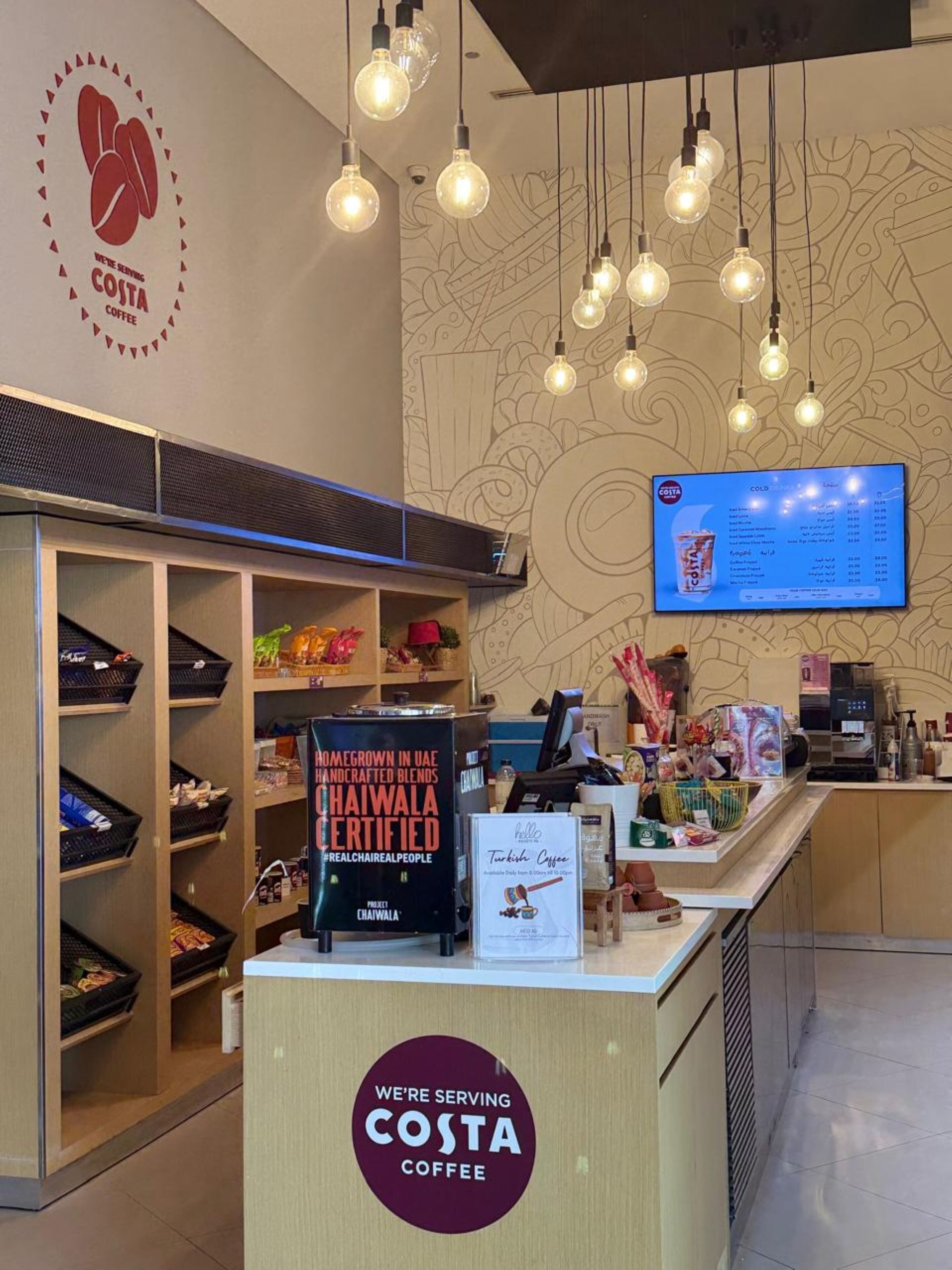 A Costa Coffee shop interior with a counter, shelves with snacks, a menu board, and hanging light bulbs.