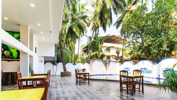 A covered outdoor dining area with wooden tables and chairs, surrounded by palm trees and a white wall with blue accents. - OYO 776 Royal Heritage (Calangute)