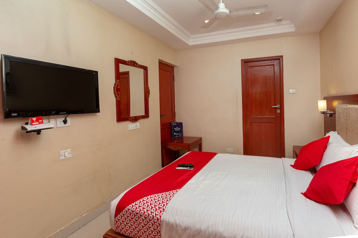 A hotel room with a flat-screen TV mounted on the wall, a bed with red and white bedding, a wooden door, and a bedside table with a lamp.