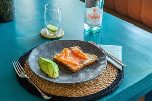 A plate with smoked salmon and avocado on toast, a fork, a knife, a glass of water, and a bottle of Acqua Panna water on a teal table.
