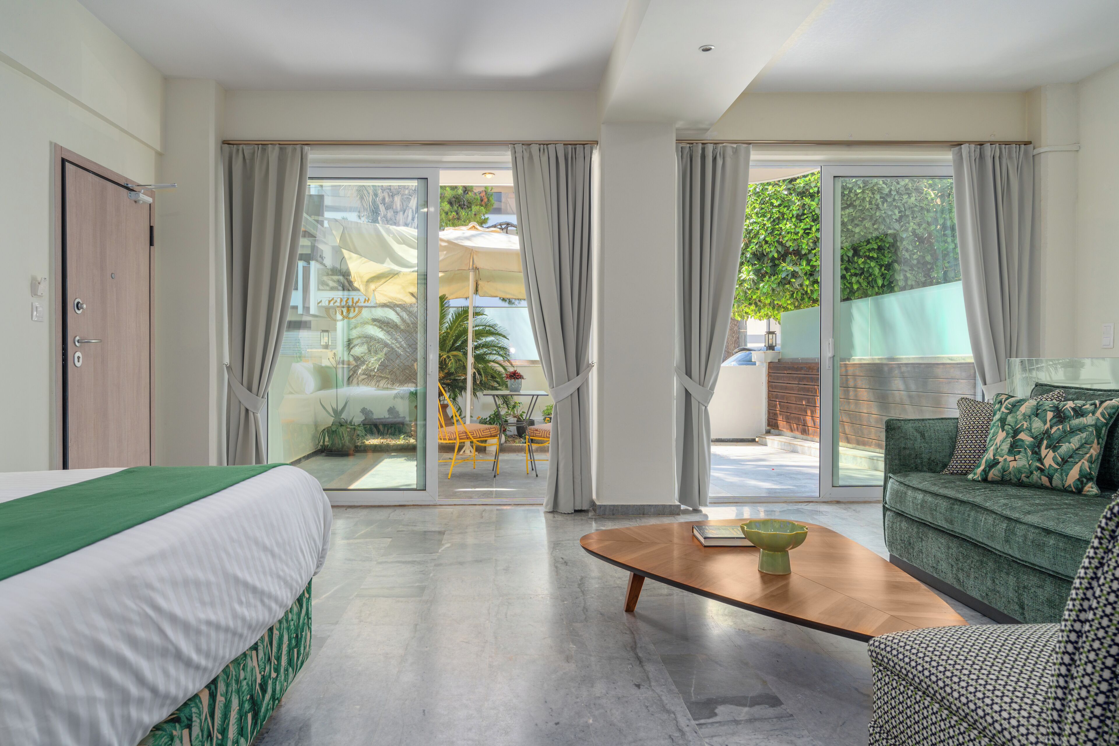 A modern hotel room with a bed, a sofa, a coffee table, and a sliding glass door leading to an outdoor area with a table and chairs.