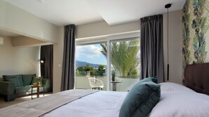 A bedroom with a large bed, a balcony with a view of palm trees and mountains, a green sofa, and a small table with a plant. - Maison 66 Riviera Hotels (Alimos)