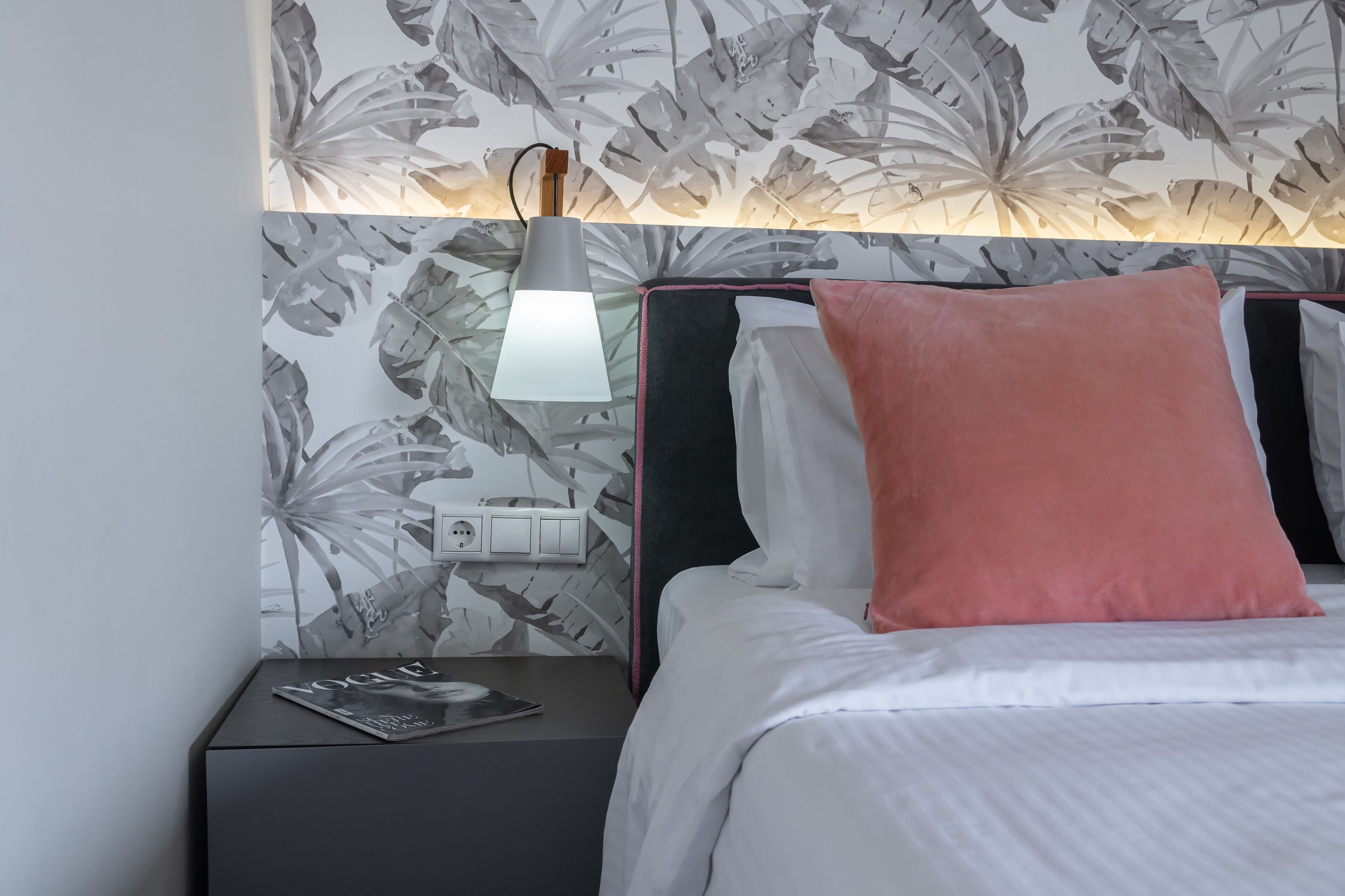 A neatly made bed with a pink pillow, a bedside lamp, and a magazine on a nightstand.