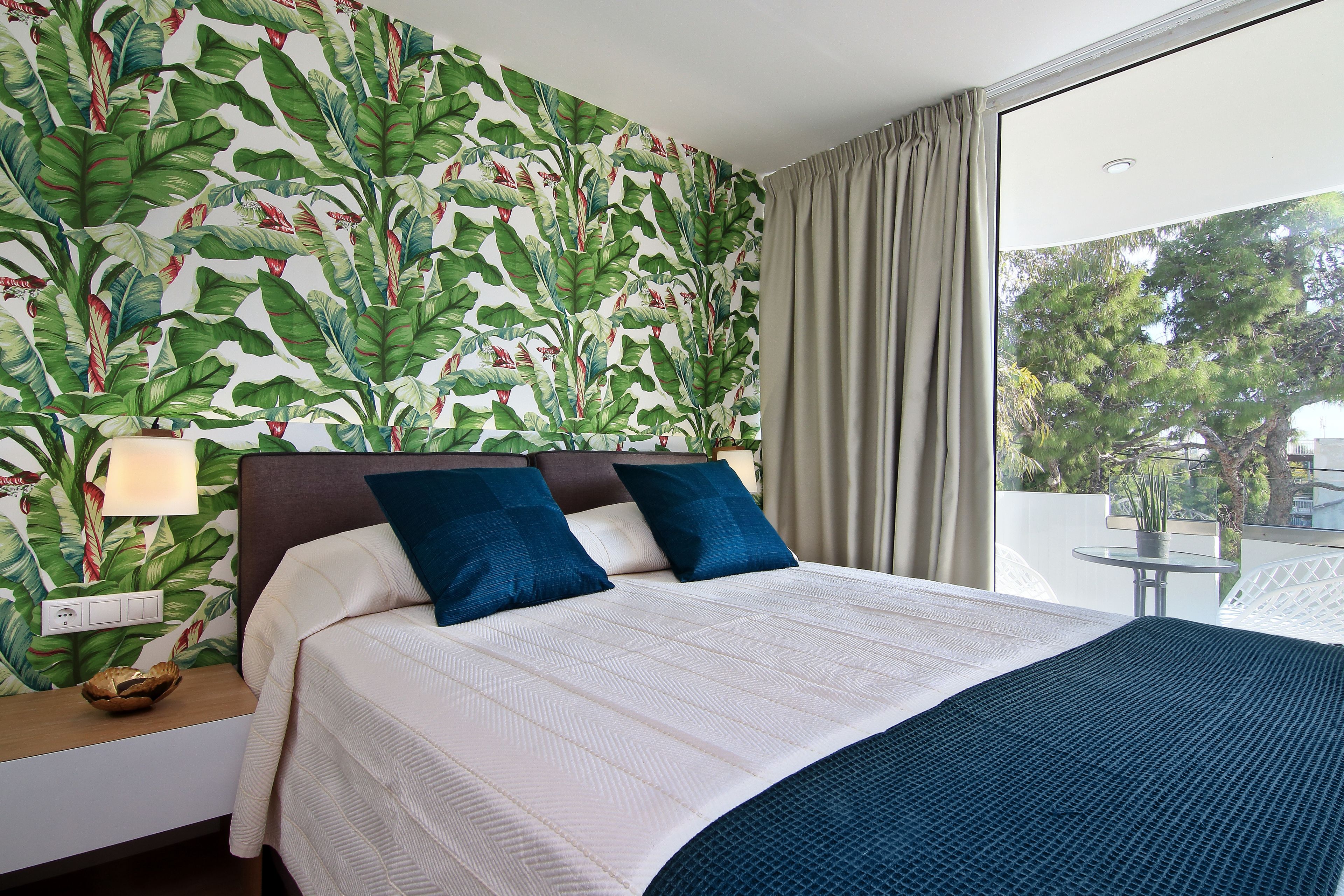 A hotel room with a large bed, a nightstand with a lamp, a view of the outdoors through a window, and a tropical wallpaper design.