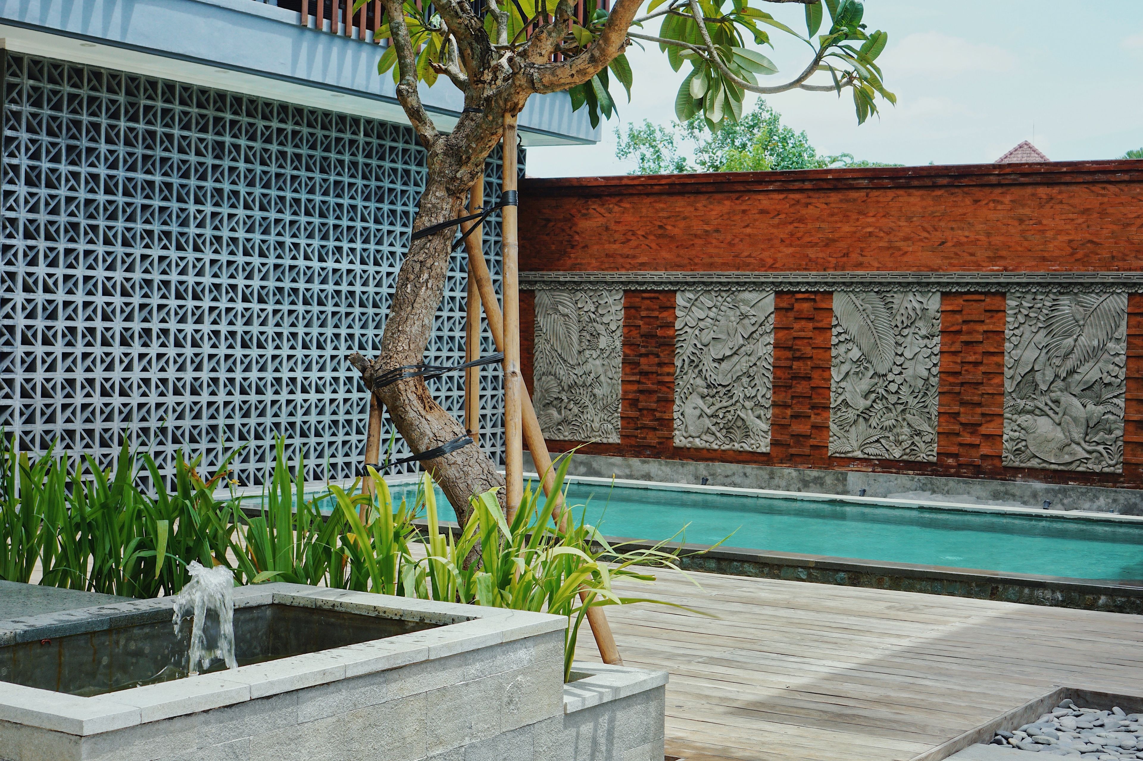 Un espace piscine agrémenté d’une terrasse en bois, d’un bac à fleurs en pierre planté, et d’un mur décoratif orné de sculptures complexes.