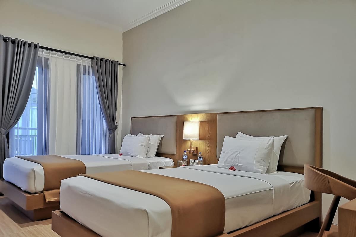 A hotel room with two beds, a wooden headboard, a bedside table with a lamp, and a window with curtains.