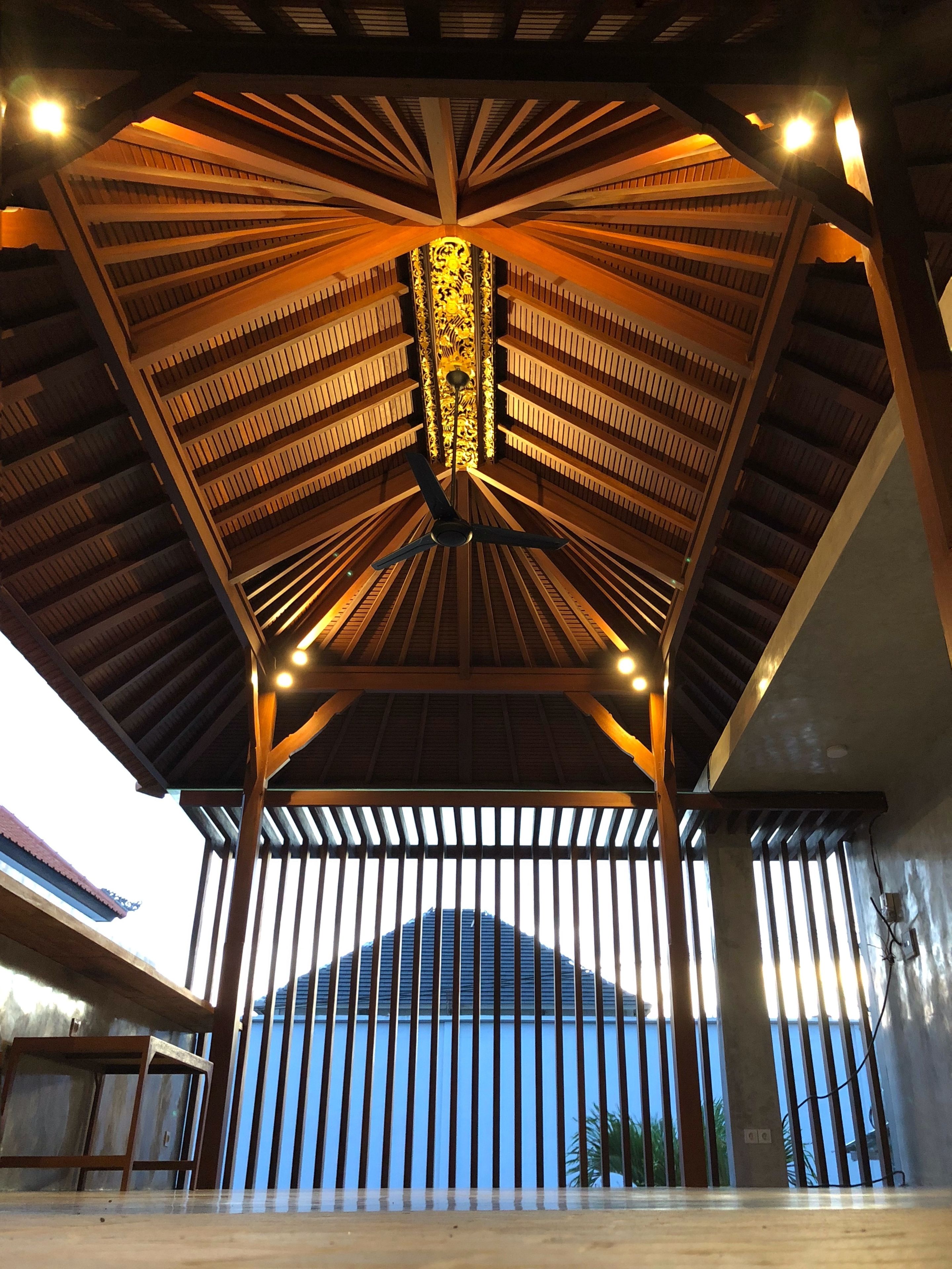 A covered outdoor area with a wooden ceiling and vertical slats, a central decorative element, and a view of a building in the background.