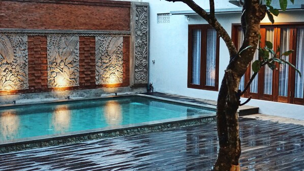 A swimming pool with intricate wall carvings, a wooden deck, and a white building with wooden shutters. - Perissos Echo Beach (Canggu)