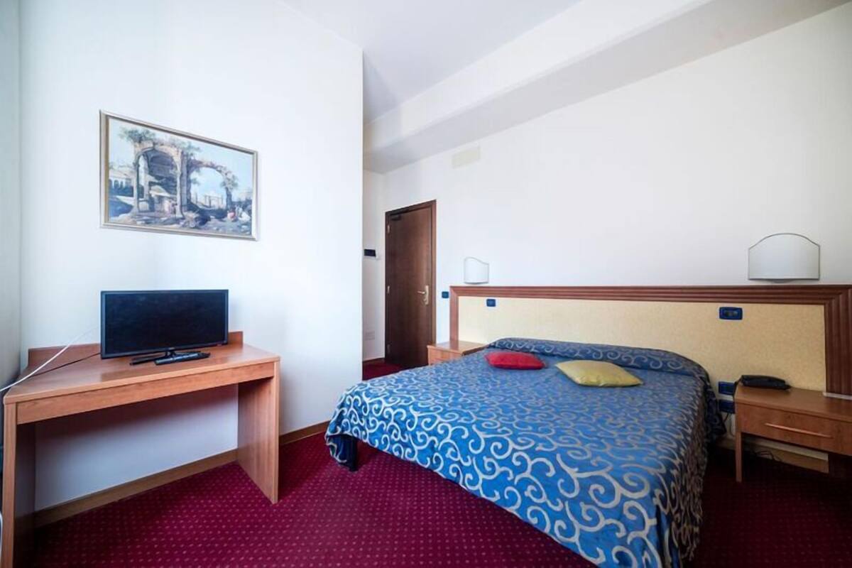A hotel room with a bed, a desk with a television, a painting on the wall, and a door.