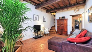 A living room with a wooden ceiling, a large plant, a TV on a stand, a sofa with red cushions, and a staircase in the background. - San Callisto - WR Apartments (Rome)