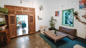 A room with a wooden door, wicker chairs, a sofa, a table with a plant, and a dreamcatcher on the wall. - Snooze Guesthouse Yogyakarta - Hostel (Yogyakarta)