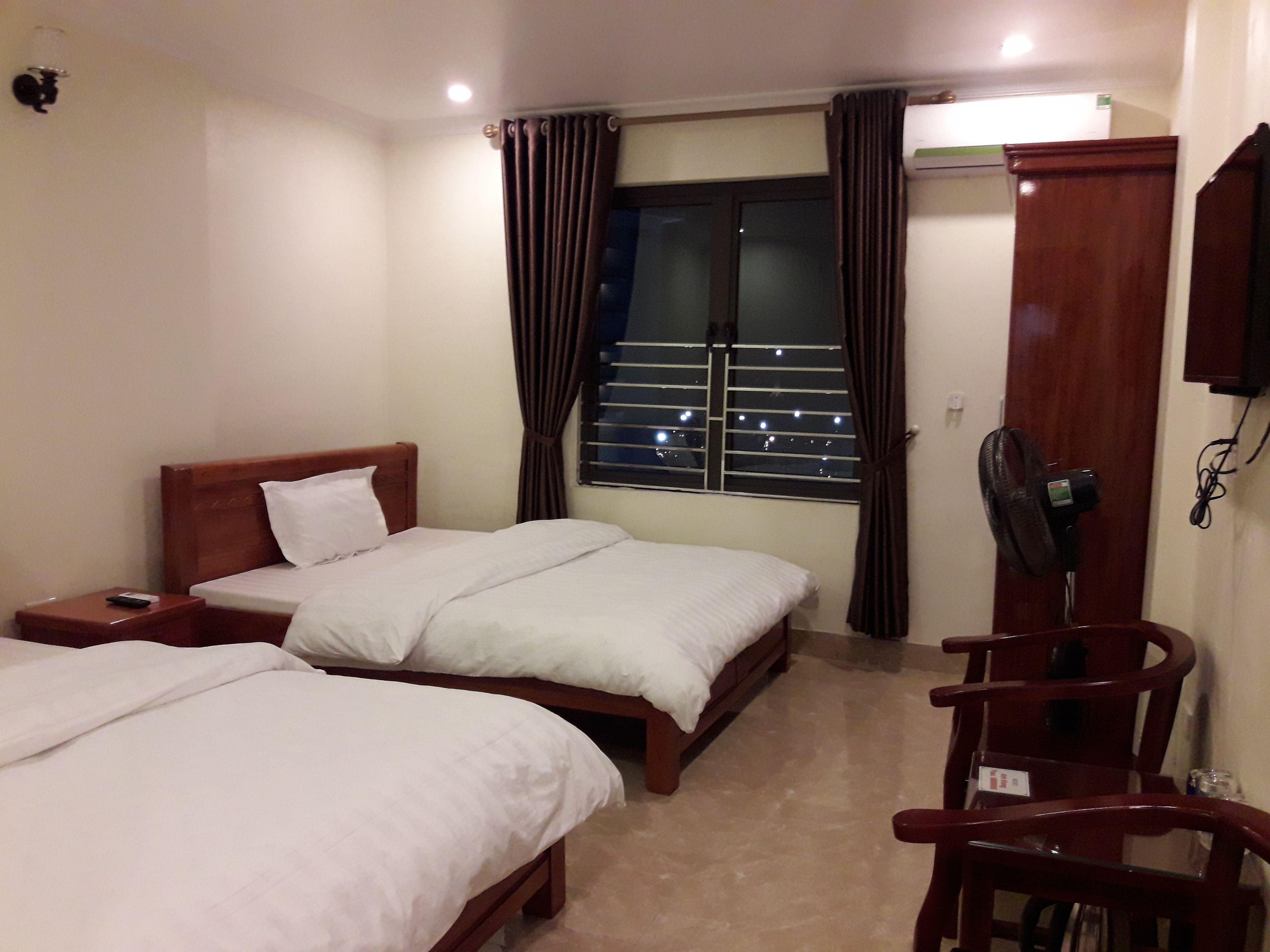 A hotel room with two single beds, a wooden nightstand, a desk with a chair, a standing fan, and a window with curtains.