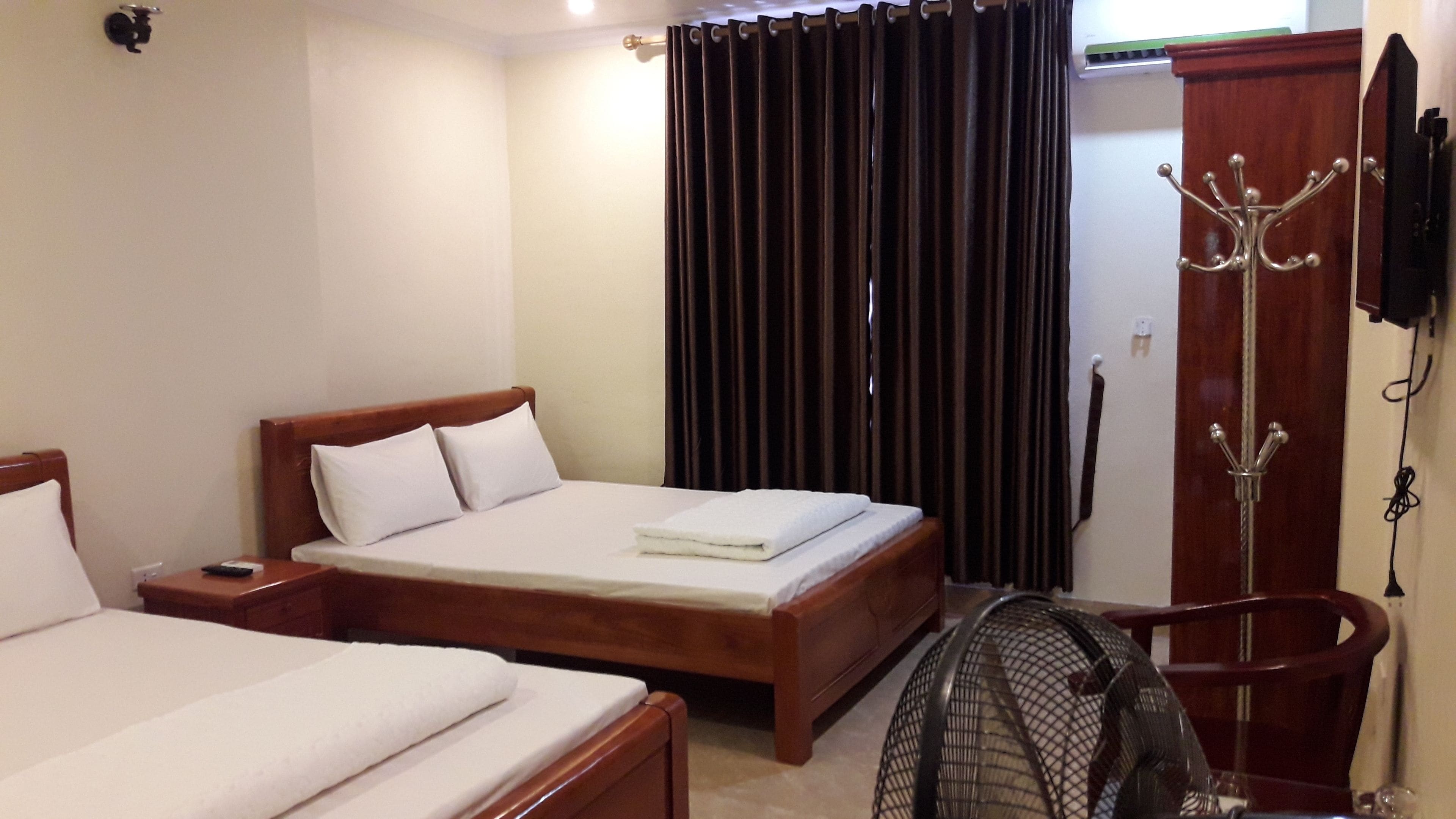 A hotel room with a double bed, a single bed, a wooden nightstand, a bedside table, a wall-mounted TV, a coat rack, and a standing fan.
