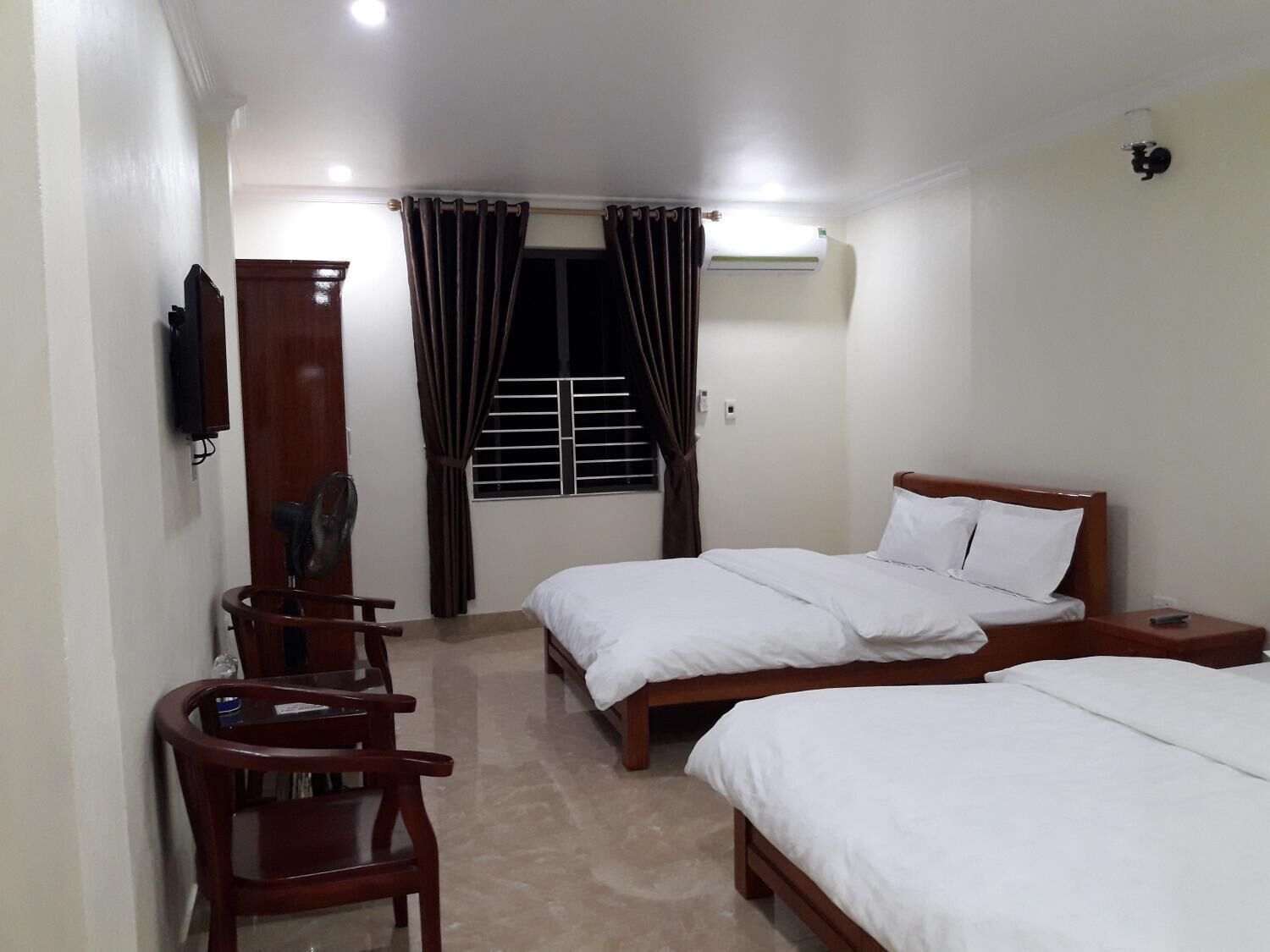 A hotel room with two beds, a wooden chair, a TV mounted on the wall, and a window with curtains.