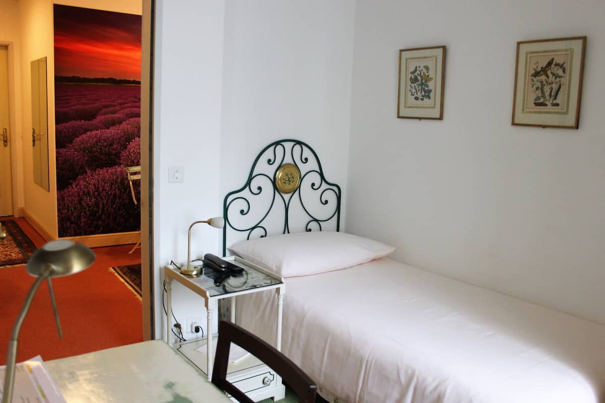 A small, neatly arranged bedroom with a single bed, a bedside table with a lamp, and a wall-mounted picture frame.