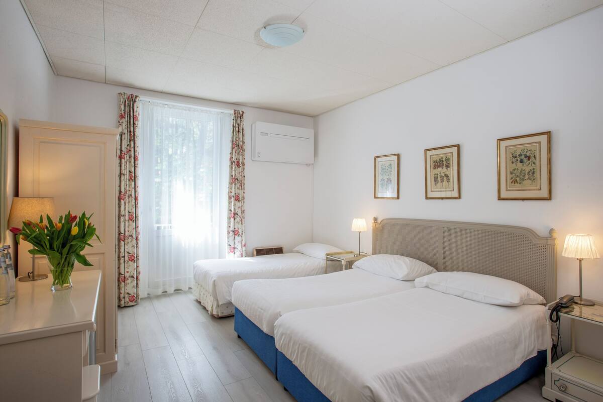 A hotel room with two beds, a desk with a lamp, a vase of flowers, and two framed pictures on the wall.